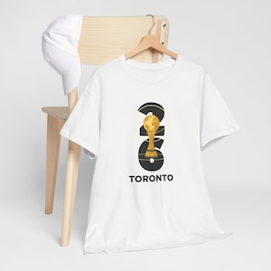 May include: White t-shirt with a graphic design featuring the number "246" in black, with a gold trophy and globe design. The word "TORONTO" is printed below the graphic in black. A white baseball cap hangs on a wooden chair.