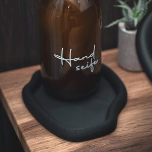 May include: A brown glass bottle labelled "Hand seife" in white script sits on a black, organically shaped tray. The tray rests on a wooden surface, with a small potted plant in the background.