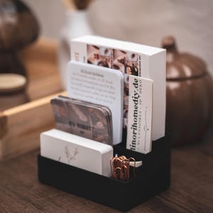 Peut inclure: Un organiseur de bureau noir contenant des cartes de visite, des brochures et des trombones. L'organisateur comporte plusieurs compartiments et est fait d'un matériau semblable à du feutre. Certaines cartes comportent du texte, dont "linorihomediy.de".