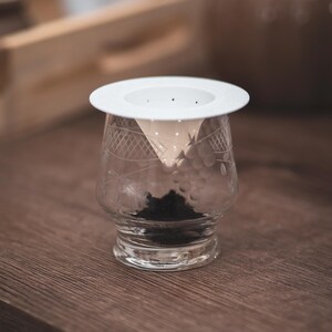 May include: A clear glass tea cup with a white tea strainer. The cup is filled with loose tea leaves. The glass has an etched design. The tea cup is on a wooden surface.