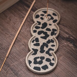 Puede incluir: Un porta incienso de cerámica beige con un diseño de estampado de leopardo negro. El soporte tiene la forma de tres círculos conectados. Un palito de incienso está insertado en el círculo superior. Un palito de incienso de madera descansa sobre la superficie de madera.