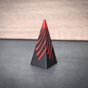 May include: A black pyramid-shaped object with red flame-like designs. The top of the pyramid is red. The object is on a gray surface with a wooden background. The design is a modern, geometric shape.