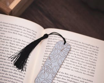 Op de afbeelding: Lichtblauw bladwijzer met een gouden bladerenpatroon en een zwarte kwast. De bladwijzer ligt op een open boek met tekst.