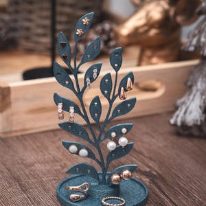 Puede incluir: Un expositor de pendientes de color verde azulado en forma de rama con hojas, que muestra varios pendientes. El expositor está sobre una superficie de madera, con anillos en la base. Los pendientes incluyen tachuelas, aros y estilos colgantes en tonos dorados y plateados.