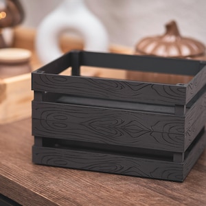 May include: A dark gray wooden crate with a wood grain pattern. The crate is rectangular and has horizontal slats. The crate is sitting on a wooden surface. Other decorative items are in the background.