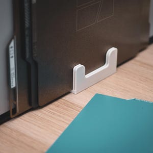 Könnte beinhalten: Ein weißer, U-förmiger Schallplattenhalter, der einen Stapel Schallplatten trägt. Der Halter steht auf einer Holzoberfläche, mit einer türkis farbenen Schallplattenhülle im Vordergrund. Die Schallplatten sind dunkelbraun.