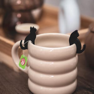Puede incluir: Una taza de color crema con un diseño acanalado, con dos sujetadores de bolsitas de té en forma de gato negro. Una etiqueta verde está adherida a la taza. La taza está sobre una superficie de madera, con objetos borrosos en el fondo.