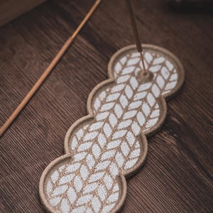 May include: A beige ceramic incense holder with a white leaf pattern. The holder is in the shape of three connected circles. An incense stick is inserted into the holder. The holder sits on a wooden surface.