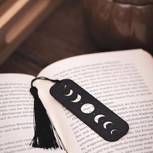 May include: Black leather bookmark with a crescent moon phase design. The bookmark is rectangular with rounded edges and a black tassel. It is placed in an open book with text.
