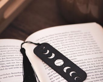 Op de afbeelding: Zwart leren boekenlegger met een maancyclus ontwerp. De boekenlegger is rechthoekig met afgeronde randen en een zwarte kwast. Het ligt in een open boek met tekst.