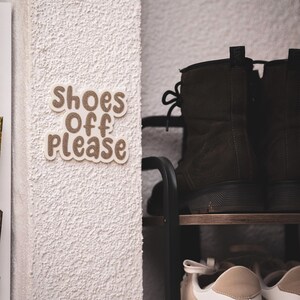 Könnte beinhalten: Eine strukturierte weiße Wand mit einem Schild mit der Aufschrift "Shoes Off Please" in braunen Buchstaben. Im Hintergrund ist ein Schuhregal mit braunen Stiefeln und beigen Turnschuhen zu sehen.