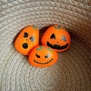 May include: Three hand-painted orange rocks with black jack-o'-lantern faces. The rocks are in a woven basket. The pumpkins have different expressions, ideal for Halloween decorations.