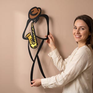 May include: A black metal wall art piece depicting a stylized figure wearing a brown hat and playing a gold saxophone. The design features clean, continuous lines forming the silhouette of a musician, with the instrument highlighted in yellow.