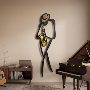 May include: A wall-mounted metal art piece depicting a stylized figure wearing a hat and playing a gold-colored saxophone. The figure is outlined in black, with a tan-colored hat and a bright gold saxophone, creating a minimalist musical silhouette.