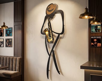 May include: A large, black, minimalist metal wall art piece depicting a stylized figure wearing a hat and playing a gold-colored saxophone. The sculpture is mounted on a light-colored wall, featuring clean, continuous lines that form the silhouette.
