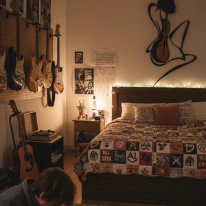 May include: A bedroom with a musical theme, featuring guitars hanging on the wall, a bed with a patchwork quilt, and a person playing a guitar on the floor. The room is dimly lit with warm lighting.