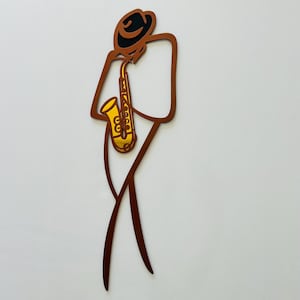 May include: A wall art piece featuring a stylized, minimalist outline of a musician wearing a hat and playing a gold-colored saxophone. The figure is constructed from thin, dark brown metal lines, creating a tall, slender silhouette against a plain background.
