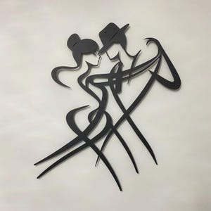 Puede incluir: Una pieza de arte mural de metal negro que presenta la silueta estilizada de dos figuras bailando. El diseño muestra una figura con un moño y otra con un sombrero fedora, conectadas por líneas fluidas y curvas que forman sus cuerpos.