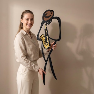 May include: A large, flat, black metal wall art piece depicting a stylized figure wearing a brown hat and playing a gold-colored saxophone. The design features bold, continuous black lines forming the silhouette of a musician in a standing pose.