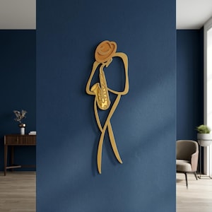 May include: A gold-colored wall art piece depicting a stylized figure wearing a hat and playing a saxophone. The design is composed of continuous, flowing lines that form the silhouette of a musician, with a detailed saxophone at the center.