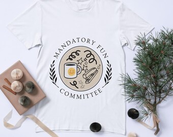 Puede incluir: Camiseta blanca con un diseño circular que dice "MANDATORY FUN COMMITTEE". El diseño incluye una taza de café con una carita sonriente, estrellas, una grapadora y remolinos decorativos, enmarcado por una corona de laurel.