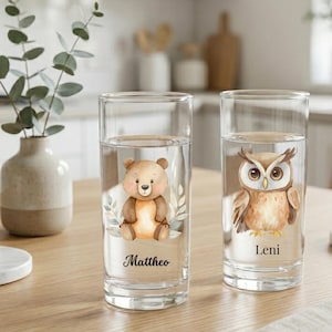 Könnte beinhalten: Zwei transparente Trinkgläser mit Wasser, jeweils mit einer Illustration verziert. Ein Glas zeigt einen Bären mit dem Namen "Mattheo", das andere eine Eule mit "Leni". Die Gläser stehen auf einem Holztisch.