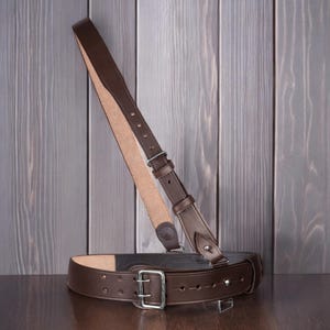 May include: A brown leather belt and shoulder strap set. The belt has a silver-coloured buckle and adjustment holes. The shoulder strap is attached with a similar buckle and is designed to be worn over the shoulder.