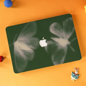 Op de afbeelding: Een donkergroene laptophoes met een wit Apple-logo en een ontwerp van twee grote, abstracte witte vlinder- of motachtige vormen. De hoes is rechthoekig.