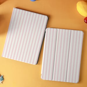 May include: Two tablet cases with a pink and white striped pattern. One case is open, revealing the interior, while the other is closed. The cases are on an orange surface with a blue ball, a lemon, and cherries.