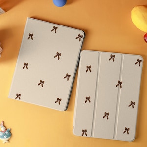 Peut inclure: Deux étuis de tablette beiges avec un motif répété de nœuds papillon bruns. Un étui est ouvert, l'autre est fermé. Les étuis sont sur une surface jaune, avec une boule bleue et des fruits décoratifs à proximité.