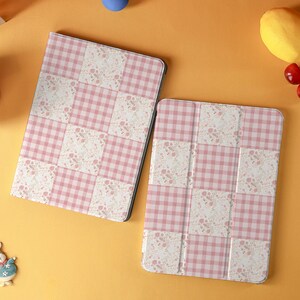 May include: Two tablet cases with a pink and white gingham pattern and floral accents. The cases are rectangular and have a folding cover. The background is a solid orange color with a few decorative items.