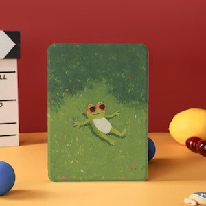 以下が含まれることがあります： サングラスをかけたカエルの漫画が草の上でリラックスしている、緑色のタブレットケース。ケースは緑色の背景に塗装されたデザインが施されています。青い球体、レモン、サクランボが背景にあります。