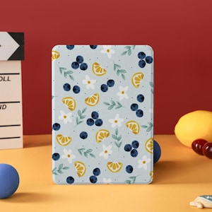 以下が含まれることがあります： ブルーベリー、レモンスライス、白い花、緑の葉の模様が入った、水色のタブレットケース。長方形のケースは赤い背景に立てられています。