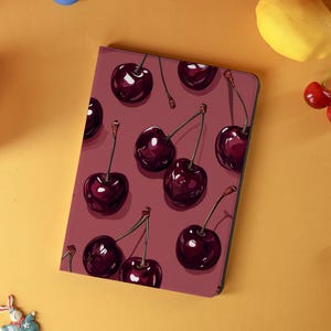 May include: A tablet case with a burgundy background featuring a pattern of dark red cherries with green stems. The cherries are evenly spaced across the surface of the case. The case is rectangular with rounded corners.