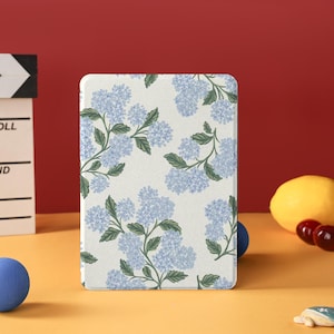 May include: A white tablet case featuring a floral pattern of light blue hydrangea blooms and green leaves. The case is rectangular with rounded corners. The background is a mix of yellow and red.