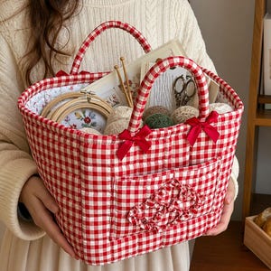 May include: A red and white gingham patterned craft basket with a quilted design. The basket has two handles and is filled with yarn, embroidery hoops, and other craft supplies. Red bows adorn the front and sides.