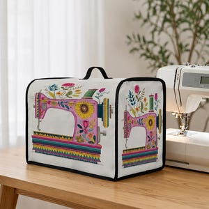 Puede incluir: Una funda blanca para máquina de coser con asa y ribete negros. La funda presenta un diseño de máquina de coser rosa con detalles florales y de girasoles. Una pila de tela colorida está en la base del diseño.
