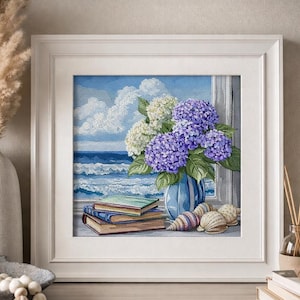 Puede incluir: Obra de arte enmarcada que representa una escena costera con un jarrón azul que contiene hortensias moradas y blancas. Libros y conchas marinas están en un alféizar con vistas al océano. La obra está en un marco blanco.