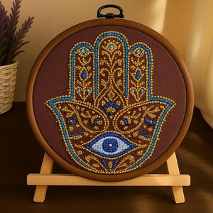 以下が含まれることがあります： 茶色の丸い刺繍フレームで、ハムサハンドのデザインが特徴です。手は青と金のビーズで装飾され、中央には目があります。この作品は小さな木製のイーゼルに飾られています。