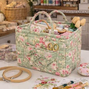 May include: A floral patterned craft organiser in green and pink, filled with sewing supplies. The quilted fabric organiser has handles and a front pocket with gold scissors. Embroidery hoops, a pincushion, and floral patterns are also visible.
