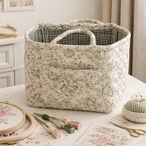 Puede incluir: Una cesta de almacenamiento de tela acolchada con un estampado floral toile de Jouy en crema y gris en el exterior y un forro de cuadros vichy en blanco y negro. La cesta tiene dos asas de tela y un bolsillo frontal rectangular con un lazo.
