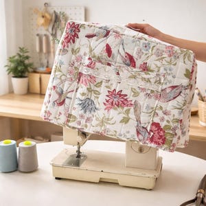 May include: A floral quilted sewing machine cover with a white background and a pattern of pink, red, and blue flowers and birds. The cover has a pocket with lace trim. The sewing machine is off-white.