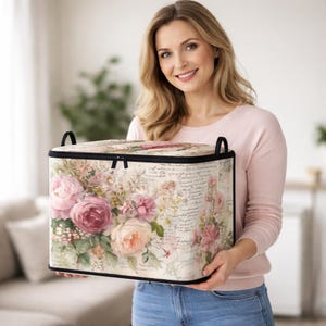 May include: A rectangular storage bin with a floral pattern featuring pink and cream roses against a background of vintage-style cursive script. The bin has a black zippered lid, black piping along the edges, and two black fabric handles on top.