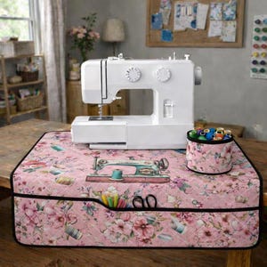 May include: A white sewing machine on a pink floral quilted cover. The cover has a vintage sewing machine illustration, spools of thread, and scissors. A matching thread holder is beside the machine. The scene is in a well-lit room.