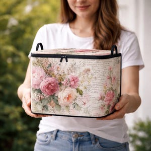 May include: A rectangular storage box with a zippered lid and two black side handles. The exterior features a cream-colored background with a vintage-style print of pink and white roses, green foliage, and faint black cursive script text.