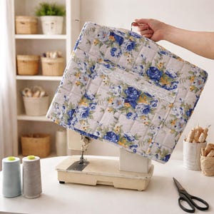 Può includere: Una copertura trapuntata per macchina da cucire con un motivo floreale di rose blu, foglie verdi e accenti gialli su sfondo grigio chiaro. La copertura presenta una tasca frontale rifinita con pizzo bianco ed è progettata per la macchina.