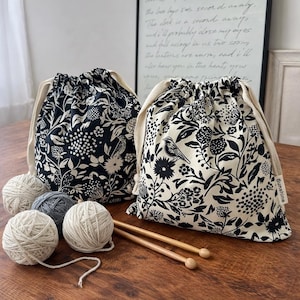 Pode incluir: Duas bolsas com cordão com um padrão floral e de pássaros em preto e branco. Uma bolsa tem fundo preto com estampa branca, a outra fundo branco com estampa preta. Inclui quatro novelos de lã e duas agulhas de tricô de madeira.