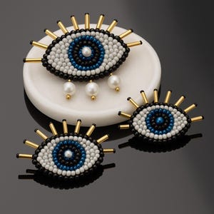 Könnte beinhalten: Drei perlenbesetzte, augenförmige Broschen. Die Augen haben eine blaue und weiße Iris, schwarz-weiße, perlenbesetzte Augenlider und goldfarbene Strahlen. Eine Brosche hat drei hängende Perlenakzente. Die Broschen liegen auf einer weißen Marmorschale und einer schwarzen Oberfläche.