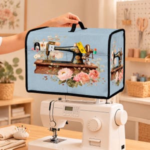 Può includere: Una copertura per macchina da cucire azzurra con una stampa raffigurante una macchina da cucire vintage circondata da fiori rosa e bianchi. La copertura presenta una maniglia nera, bordi neri e una tasca frontale con un metro a nastro giallo.