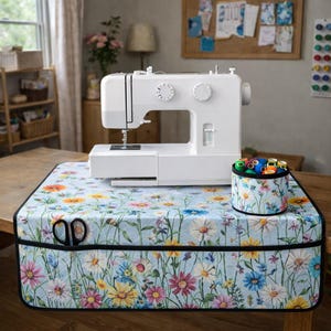 Könnte beinhalten: Eine weiße Nähmaschine steht auf einer hellblauen Hülle mit Blumenmuster. Ein passender runder Behälter enthält bunte Garnrollen. Schwarze Scheren befinden sich in einer Tasche. Die Hülle liegt auf einem Holztisch.